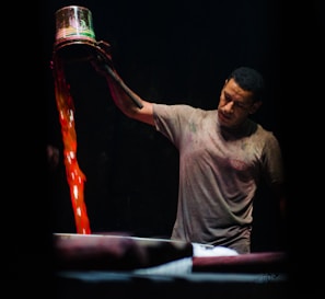 A man is pouring liquid from a metal container with vibrant red and orange hues into a large vat. He appears to be focused and intent on his task. The background is dark, highlighting both the man and the vivid colors of the liquid.