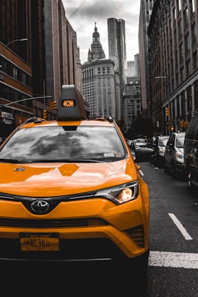 A bright yellow New York taxi is prominently displayed in the foreground, set against a backdrop of towering city buildings. The street is lined with more cars, capturing the essence of urban life. The grey, cloudy sky adds contrast to the vivid taxi, highlighting the bustling atmosphere of the city.