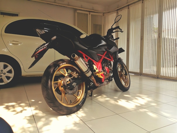 A sleek black motorcycle is parked indoors, partially illuminated by sunlight filtering through vertical blinds. The motorcycle features golden rims and a distinctive red frame, positioned next to a white car. The room has white tiled flooring and ambient natural light.