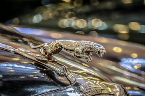 A shiny metallic jaguar hood ornament mounted on a classic car, with a background of softly blurred bokeh lights creating a luxurious and elegant atmosphere.