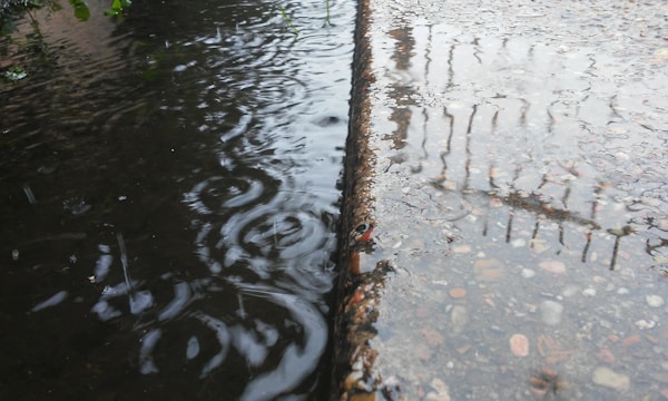 A wet surface reflects the surroundings on a rainy day. Raindrops create ripples in a puddle beside a textured concrete surface. Faint reflections of a fence or railing are visible in the water.
