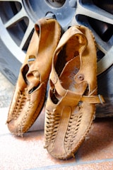 A pair of worn, handmade leather sandals with intricate weaving patterns displayed against a textured, metallic vehicle wheel. The sandals have a rustic appearance, showing signs of use and durability.