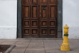 A wooden door with an intricate pattern surrounded by white walls. The base of the wall is slightly weathered, and there is a bright yellow fire hydrant on the sidewalk.