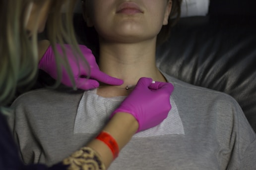 Two hands wearing pink gloves are performing a procedure or examination on the neck area of a person lying back. The person wears a grey top, and there is a white pad placed on their chest. The gloved hands focus on a small area at the base of the neck.