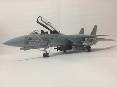 A detailed model of a military aircraft is displayed with its landing gear extended and canopy open. The aircraft features various details, including a tail number 102, and is equipped with missiles under its wings. The model is set against a neutral, light background.