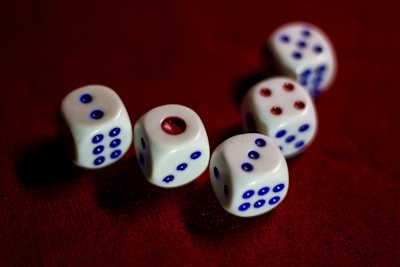 Five white dice with blue and red pips are scattered on a dark red surface. The dice are arranged in a slightly curved line, each showing a different number.