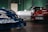 A view of three cars in a garage or showroom with green tiled walls. The cars include a blue racing car with Nissan branding partially visible in the foreground, a white car with a detailed license plate in the background, and a red car also with a visible license plate on the right. The area appears well-lit and clean, with a door labeled 'Enquiries' in the background.