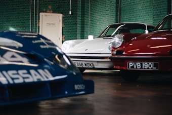 A view of three cars in a garage or showroom with green tiled walls. The cars include a blue racing car with Nissan branding partially visible in the foreground, a white car with a detailed license plate in the background, and a red car also with a visible license plate on the right. The area appears well-lit and clean, with a door labeled 'Enquiries' in the background.