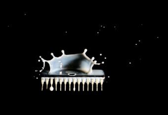 A splash of white liquid is captured in mid-air seemingly interacting with an electronic chip, set against a stark black background. The liquid creates dynamic shapes and droplets that exhibit a sense of motion and energy.