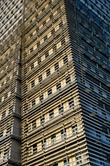 A modern building facade featuring a grid of horizontal and vertical metal beams, creating a layered, textured appearance. The facade is bathed in warm sunlight, casting shadows and highlighting the intricate structural details. Windows are visible between the beams, adding a sense of depth and complexity to the structure.