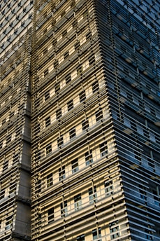 A modern building facade featuring a grid of horizontal and vertical metal beams, creating a layered, textured appearance. The facade is bathed in warm sunlight, casting shadows and highlighting the intricate structural details. Windows are visible between the beams, adding a sense of depth and complexity to the structure.