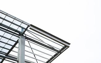 A modern architectural structure featuring steel beams and glass panels against a bright, clear sky. The design is minimalistic and emphasizes clean lines and geometric forms.