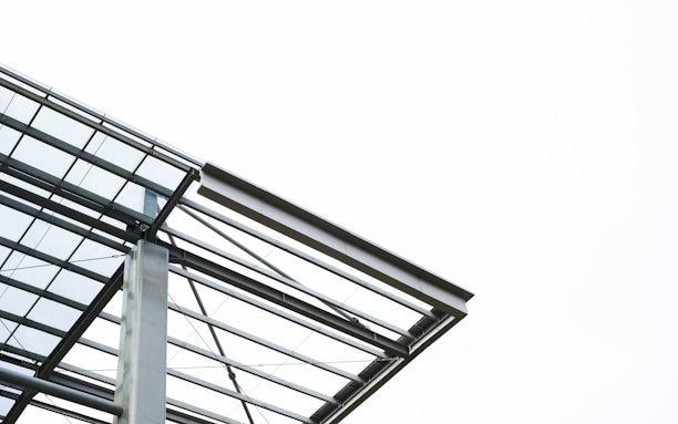 A modern architectural structure featuring steel beams and glass panels against a bright, clear sky. The design is minimalistic and emphasizes clean lines and geometric forms.