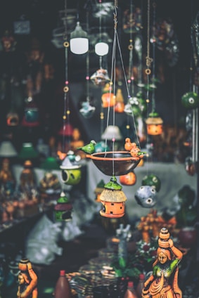 A variety of colorful handcrafted hanging decorations and birdhouses are suspended in the foreground, with traditional statues displayed in the background. The decorations feature intricate designs and vibrant hues, creating an artisanal and eclectic atmosphere.