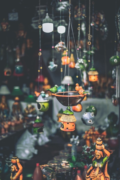 A variety of colorful handcrafted hanging decorations and birdhouses are suspended in the foreground, with traditional statues displayed in the background. The decorations feature intricate designs and vibrant hues, creating an artisanal and eclectic atmosphere.