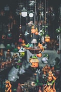 A variety of colorful handcrafted hanging decorations and birdhouses are suspended in the foreground, with traditional statues displayed in the background. The decorations feature intricate designs and vibrant hues, creating an artisanal and eclectic atmosphere.