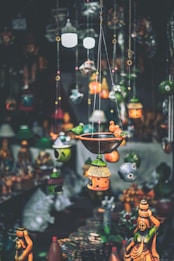 A variety of colorful handcrafted hanging decorations and birdhouses are suspended in the foreground, with traditional statues displayed in the background. The decorations feature intricate designs and vibrant hues, creating an artisanal and eclectic atmosphere.