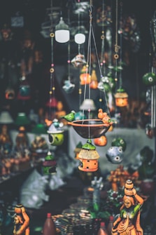 A variety of colorful handcrafted hanging decorations and birdhouses are suspended in the foreground, with traditional statues displayed in the background. The decorations feature intricate designs and vibrant hues, creating an artisanal and eclectic atmosphere.