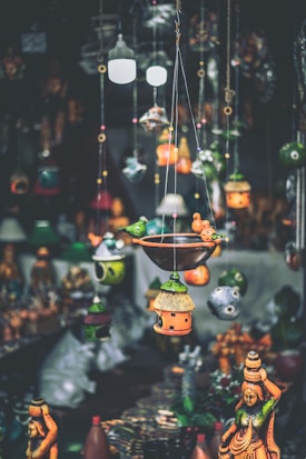 A variety of colorful handcrafted hanging decorations and birdhouses are suspended in the foreground, with traditional statues displayed in the background. The decorations feature intricate designs and vibrant hues, creating an artisanal and eclectic atmosphere.