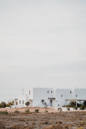 A modern, minimalist white house sits in an open, arid landscape with sparse vegetation. The building features clean lines and rectangular shapes, blending with the serene surroundings under a pale, overcast sky.
