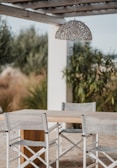 A modern outdoor dining setup with a wooden table and two white director's chairs featuring gray fabric seats and backs. Above the table hangs a decorative pendant light made of woven material. The setting is shaded by a pergola, and there are lush green plants in the background, suggesting a garden or patio environment.