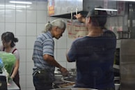 Three individuals are working in a restaurant kitchen. A man with gray hair is cooking at a stove, while another man is adjusting something near a vent. A woman stands nearby, possibly assisting with food preparation. The kitchen is enclosed with tiled walls and contains cooking equipment.