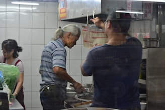 Three individuals are working in a restaurant kitchen. A man with gray hair is cooking at a stove, while another man is adjusting something near a vent. A woman stands nearby, possibly assisting with food preparation. The kitchen is enclosed with tiled walls and contains cooking equipment.