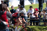 A cyclist wearing a red and black outfit with a white helmet rides a red bicycle. Several spectators, some taking photos, line a barrier on the grassy side of the road. The sun casts bright light and shadows, indicating a clear, sunny day. The cyclist is focused and appears to be in a race.