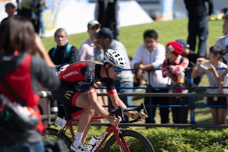 A cyclist wearing a red and black outfit with a white helmet rides a red bicycle. Several spectators, some taking photos, line a barrier on the grassy side of the road. The sun casts bright light and shadows, indicating a clear, sunny day. The cyclist is focused and appears to be in a race.