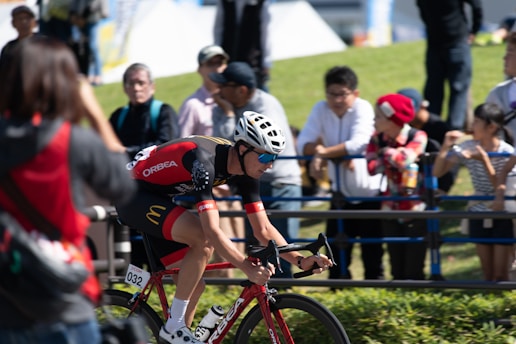 A cyclist wearing a red and black outfit with a white helmet rides a red bicycle. Several spectators, some taking photos, line a barrier on the grassy side of the road. The sun casts bright light and shadows, indicating a clear, sunny day. The cyclist is focused and appears to be in a race.