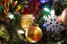 A festive display of Christmas tree decorations, featuring a Santa Claus figurine, golden and white baubles, a snowflake ornament, and various lights. The green foliage of the tree adds a vibrant background, while red berries contribute additional color.