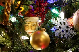 A festive display of Christmas tree decorations, featuring a Santa Claus figurine, golden and white baubles, a snowflake ornament, and various lights. The green foliage of the tree adds a vibrant background, while red berries contribute additional color.