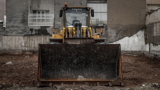 A large yellow bulldozer is parked on a dirt lot in front of a weathered building with barred windows. The machinery is centered, facing forward, and appears ready for construction work. The surrounding walls are gray and slightly damaged, contributing to an industrial or urban atmosphere.