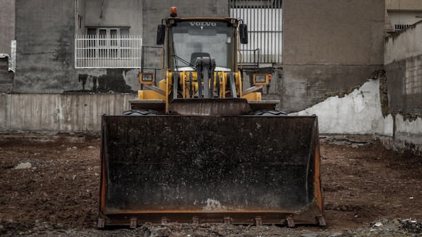 A large yellow bulldozer is parked on a dirt lot in front of a weathered building with barred windows. The machinery is centered, facing forward, and appears ready for construction work. The surrounding walls are gray and slightly damaged, contributing to an industrial or urban atmosphere.