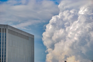 A modern high-rise building with a sleek, reflective surface stands against a backdrop of dramatic, voluminous clouds. The sky presents a gradient of blue shades, contrasting with the white and gray tones of the clouds.