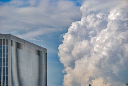 A modern high-rise building with a sleek, reflective surface stands against a backdrop of dramatic, voluminous clouds. The sky presents a gradient of blue shades, contrasting with the white and gray tones of the clouds.