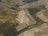 Aerial view of expansive terraced hillsides used for agriculture, featuring curving rows and patches of green vegetation amidst browner earth. Small buildings and roads are scattered throughout the landscape, with dense areas of forested greenery interspersed between the terraces.