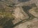 Aerial view of expansive terraced hillsides used for agriculture, featuring curving rows and patches of green vegetation amidst browner earth. Small buildings and roads are scattered throughout the landscape, with dense areas of forested greenery interspersed between the terraces.