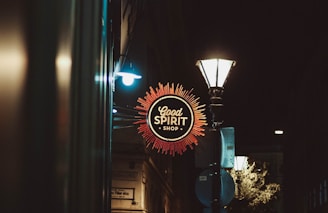 A night scene featuring a glowing neon sign for the 'Good Spirit Shop', surrounded by a decorative orange burst design. The area is dimly lit by a nearby vintage streetlamp, casting a warm light on the surrounding buildings.