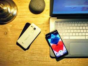 A wooden desk featuring a laptop with a visible keyboard and touchpad, a smartphone with a colorful geometric pattern on the screen, a second smartphone with 'nexus' branded on its back, a small round device resembling a smart speaker, and a metallic lamp base. The lighting is warm and casts a soft glow over the devices.