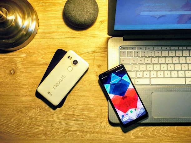 A wooden desk featuring a laptop with a visible keyboard and touchpad, a smartphone with a colorful geometric pattern on the screen, a second smartphone with 'nexus' branded on its back, a small round device resembling a smart speaker, and a metallic lamp base. The lighting is warm and casts a soft glow over the devices.