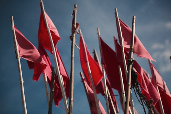 Numerous red flags are mounted on tall poles against a backdrop of a clear blue sky. The flags are in different orientations, rippled slightly, indicating a light breeze.