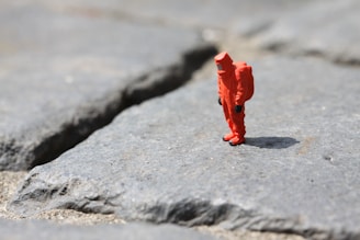 A small figurine of a person wearing an orange hazmat suit stands on a rough, rocky surface. The ground is made up of large, uneven gray stones with visible cracks.