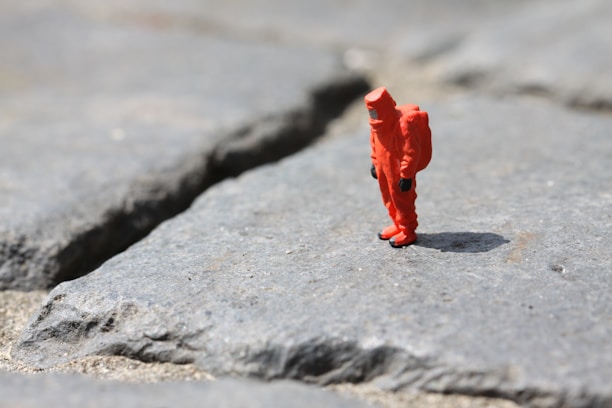 A small figurine of a person wearing an orange hazmat suit stands on a rough, rocky surface. The ground is made up of large, uneven gray stones with visible cracks.