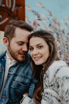 A couple is posing closely together with the woman smiling brightly at the camera. The man, wearing a denim jacket, gazes affectionately at the woman. The background features a mural with an abstract nature-inspired design.
