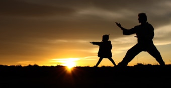 Silhouettes of two people practicing martial arts poses against a stunning sunset backdrop, with the sun peeking over the horizon and casting a warm glow.