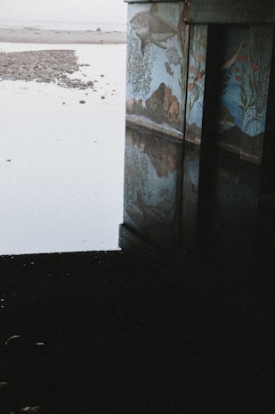 A mural depicting sea life is painted on a concrete structure near a calm, reflective water surface. The mural features various underwater creatures and plant life, creating a vibrant underwater scene. The surrounding area is serene, with a pebble-covered shore visible in the distance and sand extending toward the horizon.