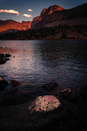 A serene mountain landscape during the golden hour, with the sun casting a warm glow over rugged peaks in the background. The foreground features a tranquil lake reflecting the sky and the surrounding rocky terrain, creating a soothing contrast between the still water and the dramatic cliffs. A few clouds float above, enhancing the peaceful atmosphere.
