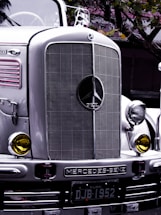 A classic Mercedes-Benz truck with a prominent front grille and headlights, bearing a vintage style. The emblem and the word 'DIESEL' are clearly visible on the grille. Yellow-tinted round headlights add to the retro aesthetic. The license plate reads 'DJB 1952', and the silver and chrome finish reflects the surrounding environment with hints of matte green leaves above.