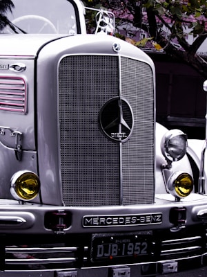 A classic Mercedes-Benz truck with a prominent front grille and headlights, bearing a vintage style. The emblem and the word 'DIESEL' are clearly visible on the grille. Yellow-tinted round headlights add to the retro aesthetic. The license plate reads 'DJB 1952', and the silver and chrome finish reflects the surrounding environment with hints of matte green leaves above.
